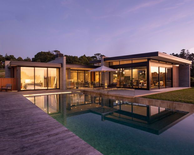 Twilight exterior view of a low-slung contemporary residence arranged around a reflective swimming pool. The home features broad floor-to-ceiling glazing, dark horizontal rooflines, warm illuminated interiors, timber decking, stone or concrete wall elements, and a covered outdoor entertaining area with an umbrella and seating. The landscaped lawn and mature trees frame the modern pavilion-like composition.