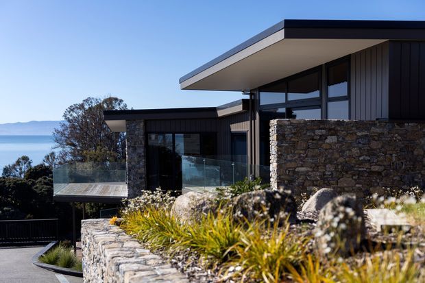 Exterior view of a contemporary multi-level home on a sloping coastal site, featuring strong horizontal flat roof planes, dark vertical cladding, extensive glazing, and layered natural stone retaining walls and facade elements. A glass balustrade borders an elevated terrace, with water and distant hills visible beyond.