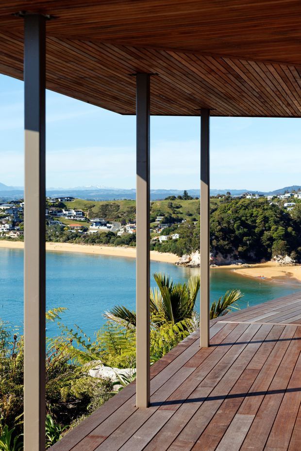 A covered timber deck or terrace overlooking a bright turquoise bay and sandy beaches. The space features dark timber decking, a richly stained timber soffit with diagonal boards, and slender dark structural posts supporting a broad cantilevered roof. Lush coastal planting frames the foreground, with residential hills and distant mountains visible beyond the water.