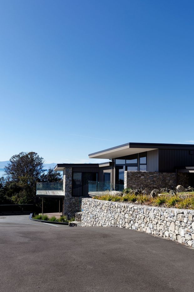 Exterior view of a contemporary single-storey home on a sloped site, featuring a low-pitched flat roof, dark vertical cladding, extensive glazing, stone-clad walls, glass balustrades, and substantial rough-stone retaining walls. A paved driveway and landscaped planting occupy the foreground, with distant hills visible beyond the property.