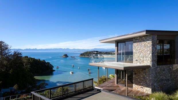 Exterior view of a modern multi-level coastal home perched above a sheltered blue-water bay. The building features a flat roof, extensive floor-to-ceiling glazing, a projecting glass-balustraded balcony, timber decking, and prominent irregular natural stone wall cladding. A dark railing and paved terrace occupy the foreground, with islands and mountains visible across the water.