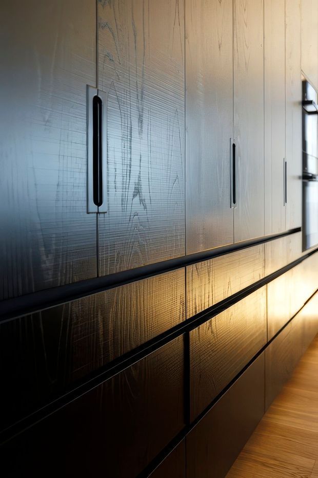 Close-up view along a sleek wall of tall built-in cabinetry with vertically textured pale panels, long black recessed handles, and darker lower cabinetry. Warm linear lighting reflects across the glossy surfaces, while a timber floor and narrow glazing are visible at the edge. The image emphasizes refined custom joinery and material detail in a contemporary interior.