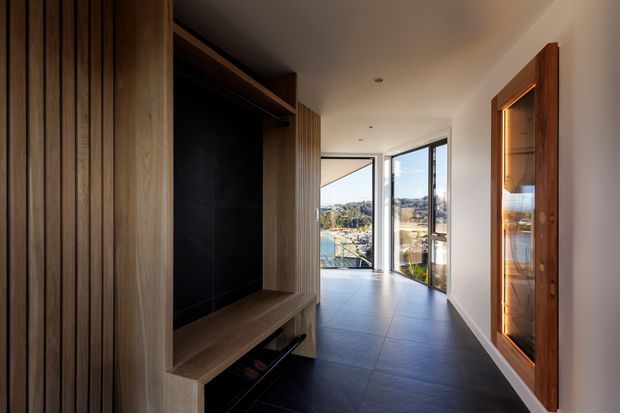 A modern residential hallway or entry passage with a built-in timber entry unit featuring vertical slatted panelling, open shelving, a bench seat and dark backing. A glazed display cabinet is integrated into the opposite wall. Large floor-to-ceiling glazing at the end of the corridor provides views toward a suburban landscape and brings in natural light. The space has dark large-format tiled flooring, white walls, warm timber joinery and a clean, refined contemporary feel.
