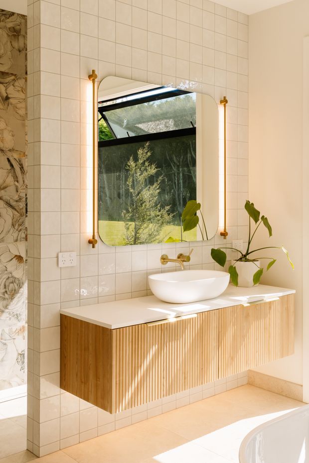 Bright contemporary bathroom vanity area with a wall-mounted fluted timber cabinet, white vessel basin, brass wall-mounted tapware, and a rounded rectangular mirror with matching brass vertical lights. The walls are clad in small glossy cream-beige square tiles, with a partial patterned feature tile wall at left. Indoor plants and a garden view through the mirror add a natural feel.