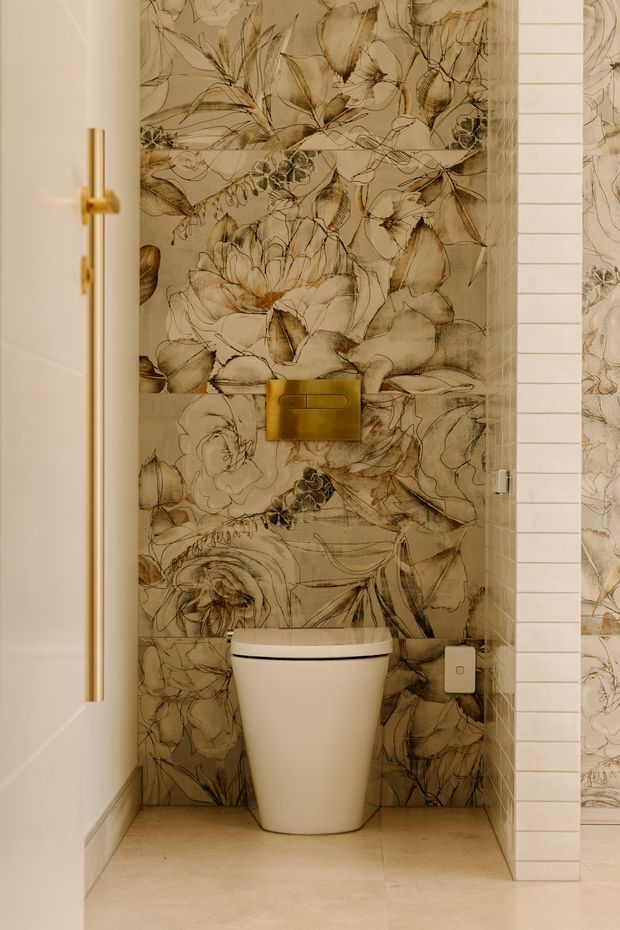 A compact contemporary bathroom WC with a wall-hung toilet set against dramatic sepia botanical wallcovering. A brushed brass flush plate and matching vertical door pull add warmth, while pale stone-look floor tiles and narrow white rectangular wall tiles frame the feature wall. A partially open white door and glass partition are visible.