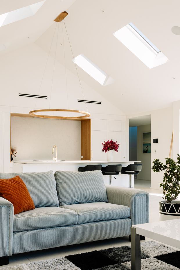 Bright open-plan living room with a pale blue ribbed sofa in the foreground, a white kitchen with timber accents and a breakfast island behind it, black bar stools, a sculptural circular pendant light, skylights, and contemporary built-in cabinetry. The room has a clean, airy aesthetic with warm timber details and contrasting black-and-white furnishings.