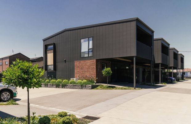 Contemporary multi-storey townhouse development with dark vertical cladding, exposed red brick ground-level walls, projecting upper volumes, covered carports, and simple landscaped planting along a broad paved driveway. The image is captured from the exterior in bright daylight.