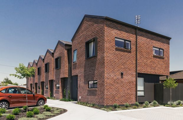 Exterior view of a contemporary multi-unit brick residential development, with a row of narrow two-storey townhouses featuring repeated gabled roof forms, dark-framed windows and doors, landscaped entry gardens, and paved driveways.