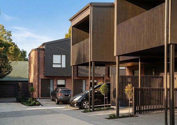 Exterior view of a contemporary multi-level townhouse development with dark timber-toned cladding, exposed red brick, vertical slatted screening, recessed balconies, and covered car parking. The buildings are arranged around a paved shared driveway or courtyard, with modern garage doors and small landscaped plantings.