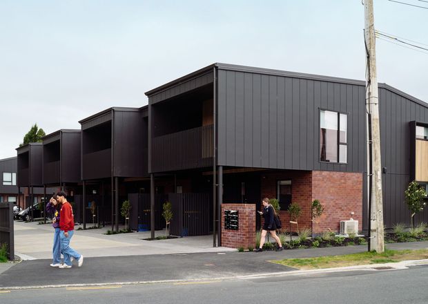 Street-level exterior view of a contemporary multi-unit residential development with dark vertical metal cladding, exposed red-brick lower walls, recessed balconies, covered ground-floor entries, and a simple flat-roofed form. Concrete paths and landscaped planting line the street frontage.