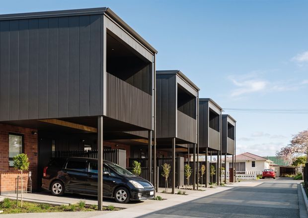 Exterior view of a contemporary multi-unit residential development with a row of narrow, stacked-looking townhouse forms. The buildings feature dark vertical cladding, projecting upper-level volumes with deeply recessed balconies, slim steel posts, ground-level carports, brick lower walls, and a shared paved access lane. Mature suburban homes are visible beyond.