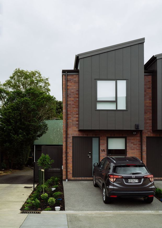 Street-facing exterior view of a contemporary attached townhouse. The facade combines red brick side walls with dark charcoal vertical metal cladding, projecting upper-level volumes, modern rectangular windows, and a dark garage and entry door. A concrete driveway, landscaped narrow garden bed, neighbouring homes, and a parked car are visible.