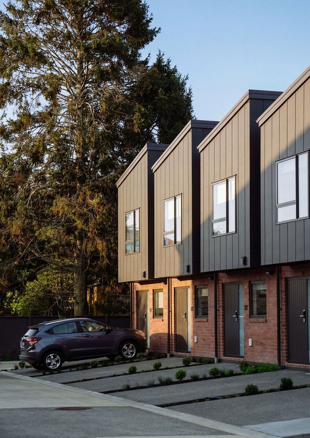 Street-level exterior view of a row of contemporary attached townhouses. The buildings feature steeply angled gabled roof forms, dark vertical metal cladding, warm brown vertical-panel cladding, exposed red-brick lower façades, tall rectangular windows, and integrated garages. A landscaped driveway with small shrubs and a parked vehicle is visible in front, with mature trees behind the development.