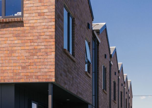 Exterior architectural view of a contemporary multi-storey building with repeating projecting brick-clad volumes, dark-framed windows, and a strong linear perspective. The facade features warm reddish-brown brickwork, dark soffits, and pitched roof elements against a clear sky.