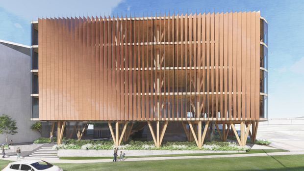 A contemporary multi-storey building facade with a rounded rectangular form, clad in closely spaced vertical timber fins that screen glazed upper levels. The structure is elevated on prominent angled timber supports, with landscaping, steps, pedestrians, and a paved forecourt at ground level. Blue sky and adjacent modern building surfaces are visible in the background.
