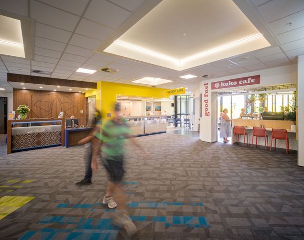 Bright contemporary commercial interior resembling a hospital or healthcare facility lobby, with a reception desk on the left, a yellow urgent care counter in the centre, and a small cafe seating area on the right. The space has a suspended acoustic tile ceiling with large illuminated recessed feature panels, patterned grey carpet, timber wall panelling, blue and yellow carpet accents, and clear wayfinding signage. Motion-blurred people are present in the foreground.