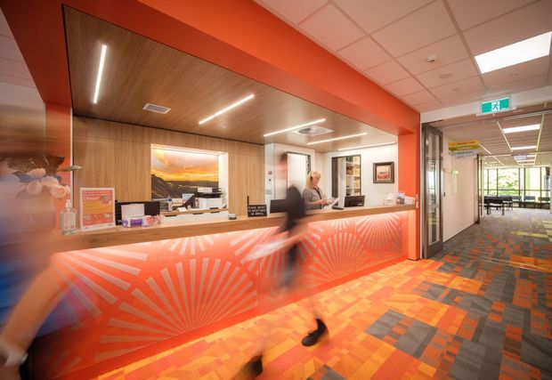 A vibrant commercial reception area with a long illuminated front desk, orange-red sunburst patterned panels, timber-look wall and ceiling finishes, framed artwork, and a patterned carpet. A corridor with glazed doors and seating extends to the right. The space appears to be a modern office, healthcare, or hospitality reception, with bright linear lighting and a bold, energetic colour scheme.
