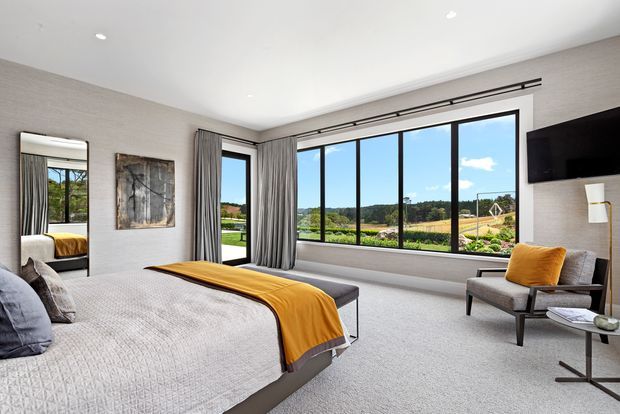 Bright contemporary bedroom with a large upholstered bed, textured light-grey bedding, and a mustard throw. Floor-to-ceiling black-framed windows and a glazed door provide expansive views of the landscaped property, while full-height grey curtains, a wall-mounted television, lounge chair, side table, artwork, and recessed ceiling lighting complete the refined interior.