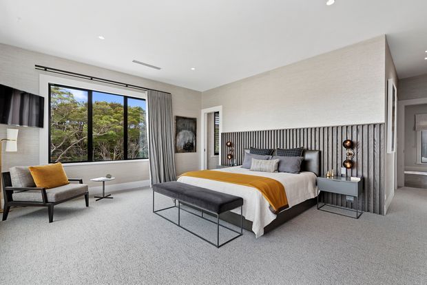 Contemporary master bedroom with a large black-framed window overlooking trees, a grey upholstered bed, mustard throw, matching bench, bedside tables, and a dramatic vertically fluted feature wall behind the bed. The room has soft neutral finishes, recessed lighting, full-height curtains, and a calm, refined atmosphere.