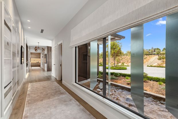 Bright, refined residential hallway with full-height built-in cabinetry on the left, a large floor-to-ceiling window overlooking landscaped grounds, pale carpet, timber-toned flooring at the edges, and contemporary recessed and pendant lighting. The corridor leads toward a warmly lit living or dining area.