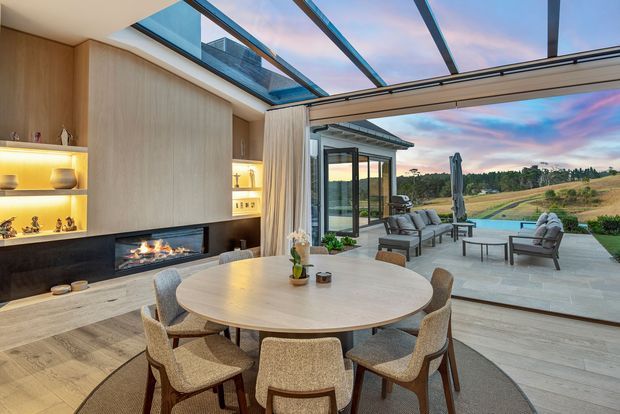 A refined open-plan dining and living space with a round timber dining table and upholstered chairs in the foreground. Built-in timber cabinetry with illuminated display shelving and a linear fireplace forms the feature wall. Full-height retractable glazing opens to a paved outdoor terrace with lounge seating and expansive landscaped countryside views, while a glazed roof brings in natural light.