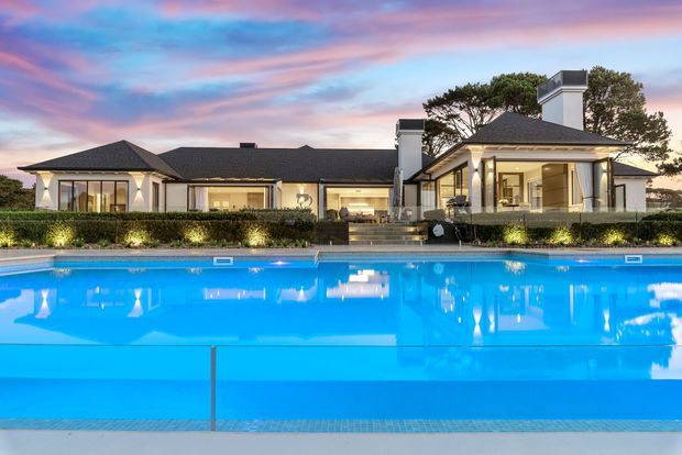 A large, elegant single-storey residence viewed across a bright swimming pool at dusk. The home has a dark hipped roof, pale rendered walls, expansive glazed openings, prominent chimneys, and a symmetrical pavilion-like composition. Landscaped hedging and outdoor lighting frame the pool, while the colourful sunset reflects in the water.