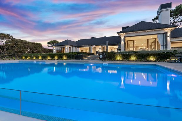 A large illuminated swimming pool dominates the foreground of a substantial single-storey residence at dusk. The home features dark hipped roofs, pale rendered walls, large glazed living areas, classical columns and prominent chimney structures, with hedging and mature trees surrounding the pool.