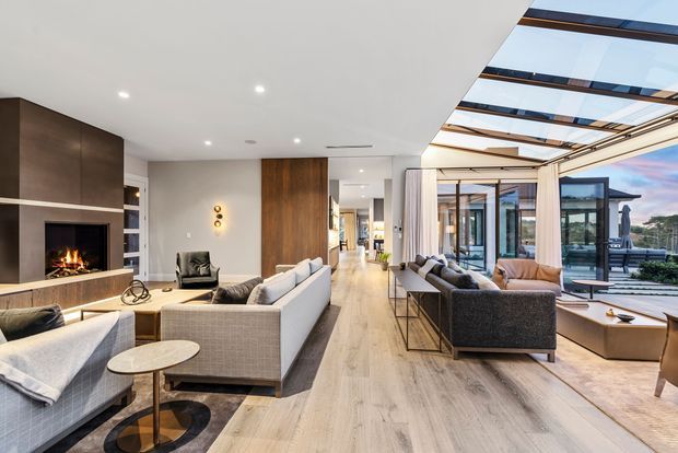 A spacious contemporary open-plan living room with pale timber flooring, a linear fireplace set into dark wood joinery, neutral upholstered sofas, and a long central circulation zone. Floor-to-ceiling glazing and a glazed roof structure create a bright connection to the outdoor terrace and water-facing landscape. The room includes refined black-framed glass doors, a low coffee table, recessed ceiling lighting, and warm timber feature panels.