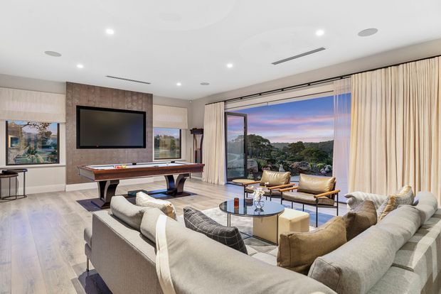 A spacious, refined living room with a large sectional sofa arranged around a round glass coffee table and ottomans. The room includes a pool table, wall-mounted television on a stone-textured feature wall, expansive glazed doors opening to a scenic outdoor view, full-height cream curtains, recessed lighting, and pale timber flooring.