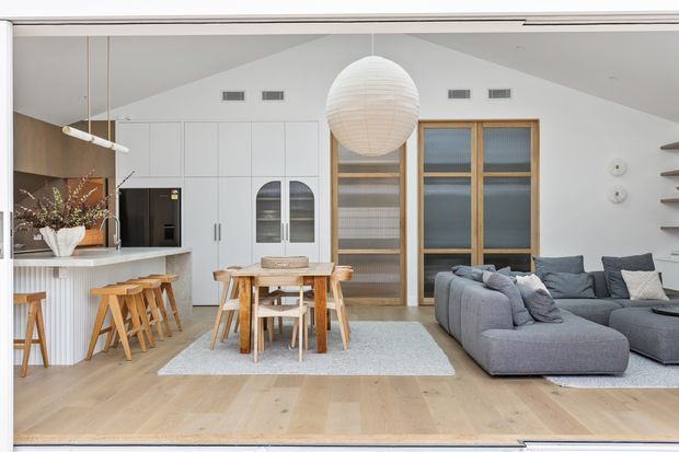 Bright open-plan interior showing a contemporary kitchen, dining area and living room. The space has pale timber flooring, white built-in cabinetry, a stone kitchen island with timber stools, a timber dining table and chairs, a large grey sectional sofa, timber-framed glazed doors, floating wall shelves and a prominent spherical pendant light.