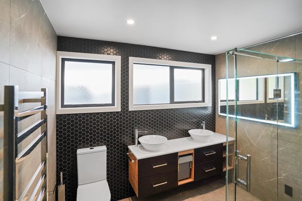 Contemporary bathroom featuring a floating dark timber vanity with two vessel basins, a white toilet, a heated towel rail, black hexagonal wall tiles, pale stone-look wall surfaces, frosted horizontal windows, and a frameless glass shower enclosure with integrated lighting.
