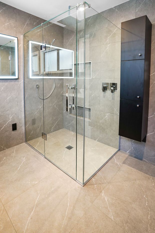 Contemporary bathroom featuring a large frameless glass walk-in shower with chrome hardware, grey-beige veined stone-look wall and floor tiles, illuminated wall mirrors, chrome shower fixtures, and a tall dark built-in storage cabinet.