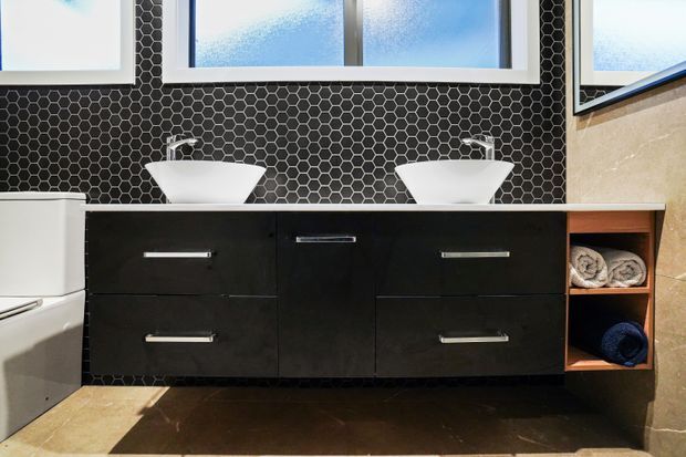 Contemporary double vanity bathroom featuring a floating black vanity with two white vessel basins, chrome tapware, open timber towel shelving, and a dramatic black hexagonal tile splashback. Frosted windows and warm stone-toned floor and wall surfaces complete the high-contrast design.