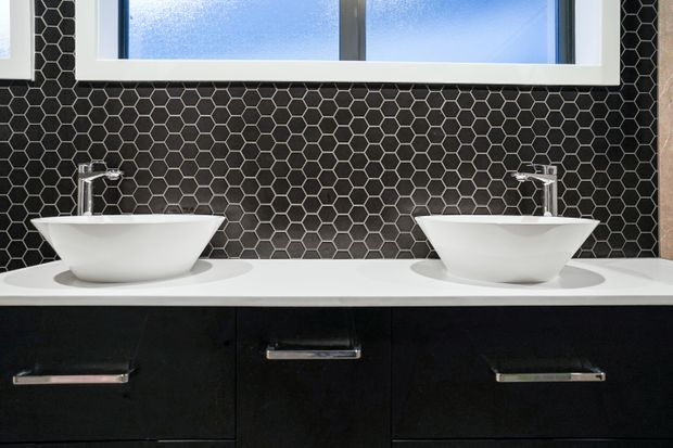 A contemporary bathroom vanity with two white vessel basins and chrome taps set on a white benchtop above dark cabinetry. The backsplash is clad in small black hexagonal tiles, with a frosted horizontal window above.