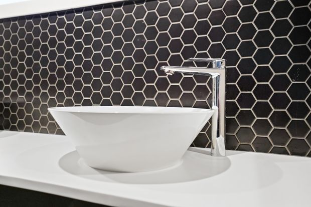 Close-up of a contemporary bathroom vanity featuring a white vessel basin on a white countertop, a chrome geometric tap, and a dark hexagonal tile splashback with light grout.