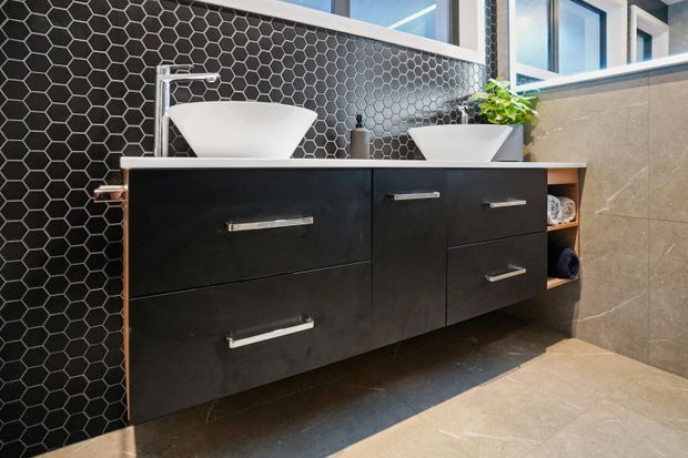Contemporary bathroom vanity with two white vessel basins set on a floating dark cabinetry unit. The wall behind features black hexagonal mosaic tiles, while the adjacent wall and floor are clad in large neutral stone-look tiles. Chrome tapware, open shelving, and high windows complete the modern fit-out.