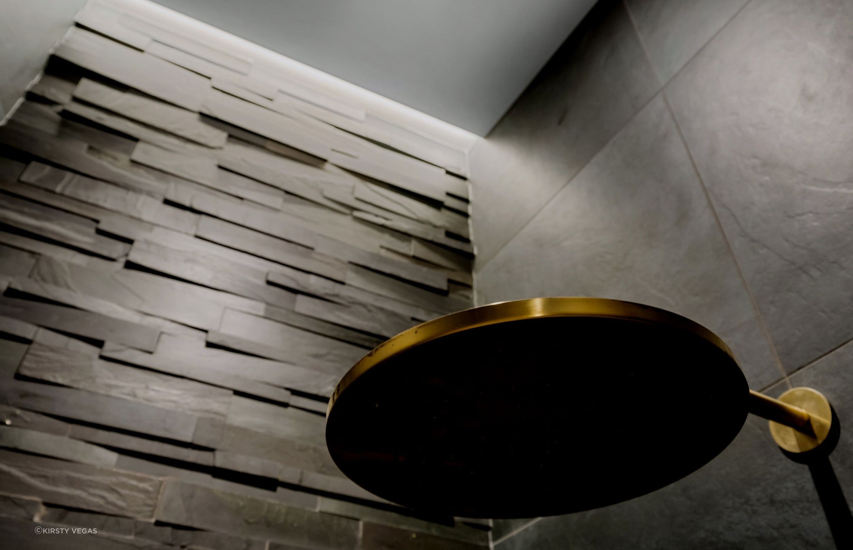 The stunning aged brass Progetto shower head creates a striking contrast against the impressive four-meter 'Island Stone' feature wall, enhancing the visual appeal of the space.