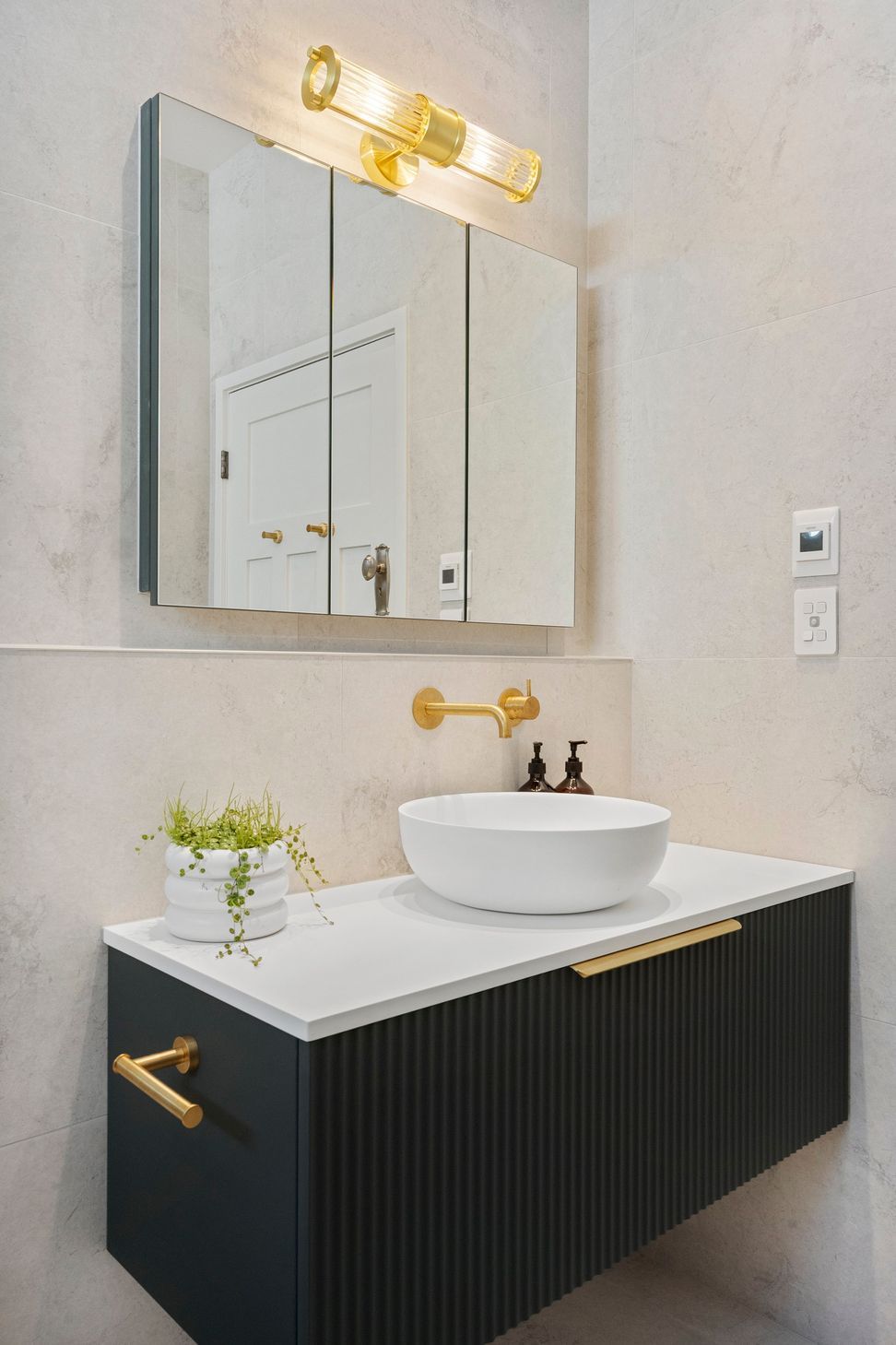 The Vista mirrored wall unit enhances the bathroom's functionality by providing additional storage and convenient power outlets, all while contributing to the sleek design. Its reflective surface amplifies natural light, creating a sense of spaciousness. Complementing this piece, the fluted glass wall lamp perfectly aligns with the fluted vanity and other brushed brass elements, creating a cohesive and harmonious aesthetic. Together, these features add both practicality and style, elevating the overall elegance of the bathroom.
