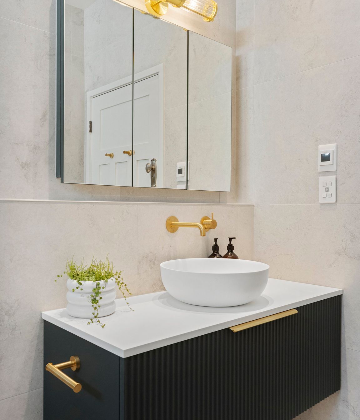 The Vista mirrored wall unit enhances the bathroom's functionality by providing additional storage and convenient power outlets, all while contributing to the sleek design. Its reflective surface amplifies natural light, creating a sense of spaciousness. Complementing this piece, the fluted glass wall lamp perfectly aligns with the fluted vanity and other brushed brass elements, creating a cohesive and harmonious aesthetic. Together, these features add both practicality and style, elevating the overall elegance of the bathroom.