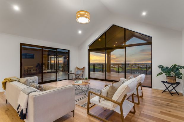 Bright contemporary living room with white walls and a warm timber floor, furnished with a light sofa, two timber-framed lounge chairs, a coffee table and a potted plant. A large gabled glazed wall opens to a balcony or terrace and provides expansive views, while dark-framed sliding glass doors connect to an adjacent space. A sculptural pendant and recessed ceiling lights add understated detail.