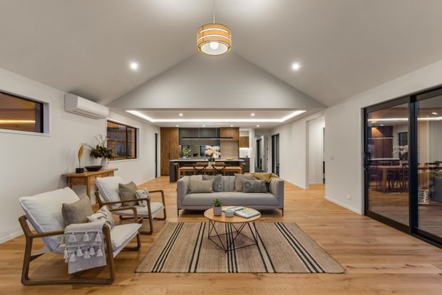 A bright, open-plan contemporary living room with a pitched ceiling and timber flooring. The room includes a grey sofa, armchairs, a low coffee table, and a striped area rug, with a kitchen and dining area visible beyond. Large black-framed glazed doors open to an adjacent space, while recessed lighting and a central pendant provide illumination. White walls and warm timber finishes create a clean, inviting atmosphere.