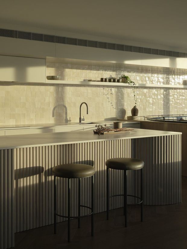 A warm, refined contemporary kitchen featuring a large island with a vertically fluted, curved-panel base, pale stone benchtop, dark bar stools, and integrated cabinetry. Glossy cream square tile forms the splashback, with open shelving, concealed rangehood ventilation, a sink, cooktop, and understated decorative styling. Strong late-afternoon light creates a calm, softly dramatic atmosphere.