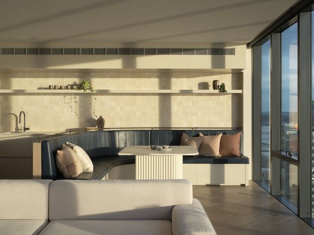A warm, contemporary open-plan living and dining area with a built-in dark blue banquette, pale upholstered sofa, ribbed pedestal dining table, and a light stone-tiled feature wall with open shelving. Full-height glazed doors or windows line the right side, bringing in abundant natural light and views beyond. The room has timber flooring, integrated dark cabinetry, and a refined neutral palette.