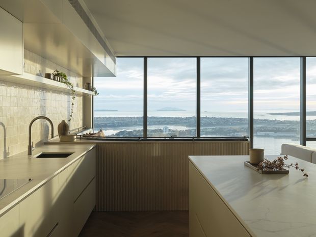 A refined contemporary kitchen with a large island and integrated cabinetry, overlooking a broad coastal or waterfront cityscape through expansive floor-to-ceiling windows. The space features pale stone surfaces, light neutral cabinetry, a dark timber floor, a ribbed timber-clad window wall, and brushed metal-framed glazing. A minimalist pendant-free ceiling and restrained styling create a calm, elevated atmosphere.
