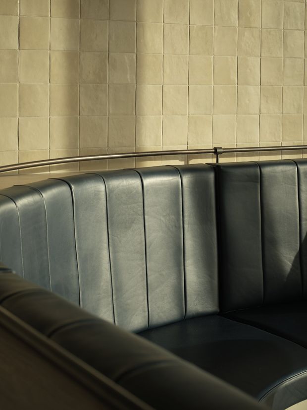 Close-up interior view of a dark upholstered booth with vertically channelled black or charcoal leather seating. A slim metal handrail runs along the top against a wall clad in warm, irregular square beige tiles. The scene has a refined, moody hospitality or restaurant atmosphere with strong directional light and shadow.