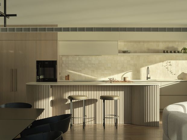 A warm, contemporary open-plan kitchen and dining area with a long island featuring vertically fluted cabinetry, integrated appliances, pale stone or tile surfaces, and a textured light backsplash. Three bar stools sit at the island, with a dining table and chairs in the foreground. Soft late-afternoon light creates strong shadows across the cabinetry and floor.