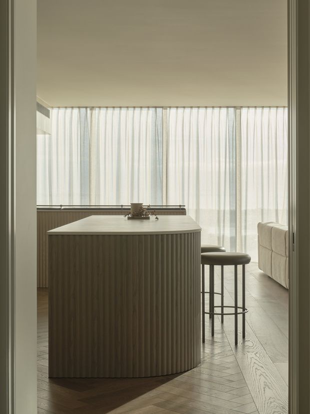 Warm, softly lit contemporary kitchen viewed through a doorway, featuring a large rounded island with vertically fluted cabinetry and a pale smooth benchtop. Two slim bar stools sit at the island, while sheer full-height curtains diffuse light across the room and a pale upholstered sofa is visible beyond. The floor is laid in a dark-to-mid timber herringbone pattern, creating a refined, calm atmosphere.
