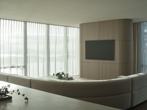 A serene contemporary living room viewed from behind a large curved sectional sofa. Floor-to-ceiling sheer curtains provide soft daylight, while a curved timber-panelled media wall with integrated television forms the main feature. A pale stone or light-coloured island-like surface occupies the foreground, with minimal greenery and decor.