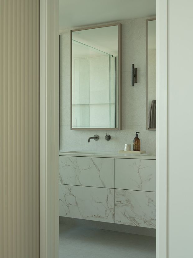 A refined contemporary bathroom viewed through a doorway, featuring a floating double vanity clad in pale white marble-look panels with grey veining, a matching light stone benchtop and splashback, wall-mounted brushed metal tapware, frameless glass shower enclosure, large mirrored cabinets, and a slim dark wall light. The room has soft neutral tones, clean lines, and a ribbed vertical wall detail at the left edge.