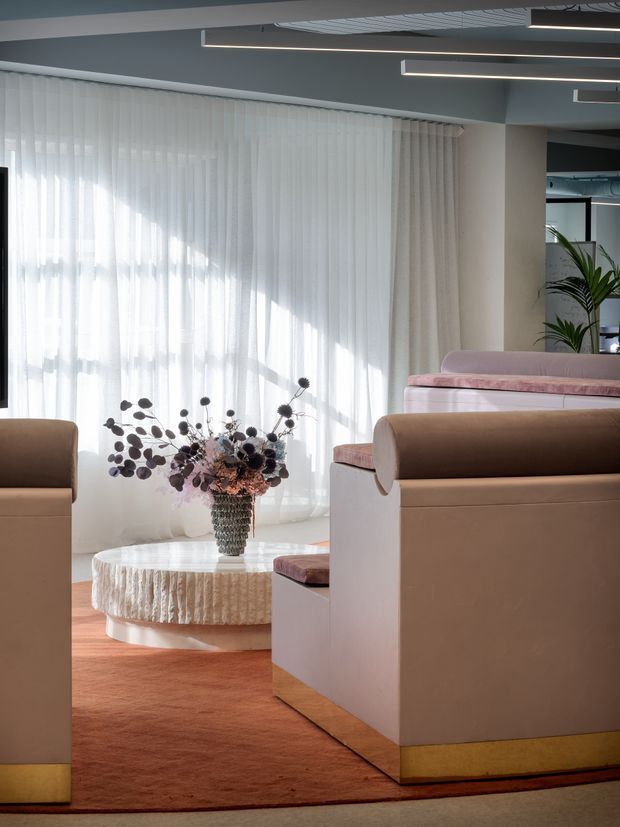 A refined contemporary lounge interior with large sheer curtains, sculptural upholstered armchairs and sofas, a round textured coffee table, and a decorative floral arrangement. Warm terracotta carpet and blush, taupe, and cream upholstery contrast with the cool grey ceiling and linear modern lighting.